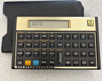 Könnte beinhalten: Ein Hewlett-Packard 12C Finanzrechner mit goldfarbener Frontplatte, schwarzen Tasten und einem Digitaldisplay, das 0.00 anzeigt. Das Gerät hat ein horizontales Layout mit zahlreichen Funktionstasten und liegt vor einem schwarzen Etui.