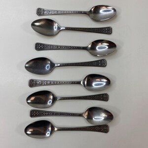 May include: An overhead shot of seven silver spoons with ornate floral and geometric designs on the handles. The spoons are arranged on a white surface, showcasing their polished, reflective bowls. The spoons are likely for serving or eating.