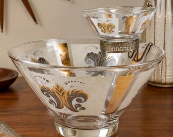 Puede incluir: Un juego de servir para aperitivos de cristal transparente que incluye un cuenco grande y uno más pequeño acoplado. Ambas piezas están decoradas con un patrón repetitivo de motivos florales estilizados en dorado y gris y paneles verticales.