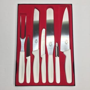 Può includere: Un set di sette coltelli e utensili da cucina in acciaio inossidabile con manici bianchi, tra cui una forchetta da intaglio, un coltello da cucina, un coltello da pane e un pelapatate, presentati in una scatola foderata di velluto rosso. Le lame hanno una finitura argentata.