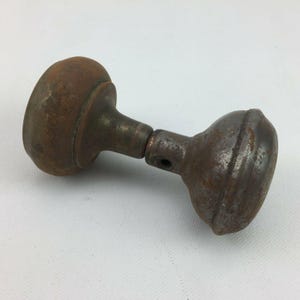 Puede incluir: Un pomo de puerta de metal vintage con un acabado desgastado y oxidado en tonos marrón y gris. El objeto consta de dos extremos redondeados y abultados conectados por un eje central cilíndrico, con una pequeña abertura circular en el cuello.