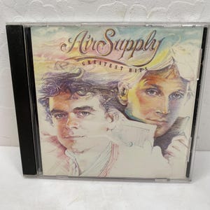 May include: A CD case with a colorful illustration of two men. The case is titled "Air Supply Greatest Hits".