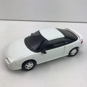 以下が含まれることがあります： 黒いフロントガラスと窓が付いた白いおもちゃの車。車にはシルバーのホイールとスポイラーが付いています。車はクーペスタイルです。
