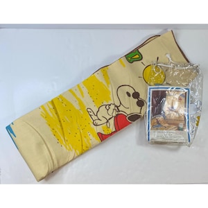 Puede incluir: Una manta Snoopy plegada, de color crema, con un diseño amarillo y rojo. La manta presenta a Snoopy y Woodstock en una pose juguetona. También se ve un artículo empaquetado con una imagen de Snoopy.