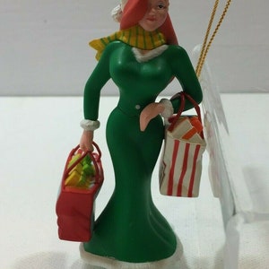 May include: A red-haired woman wearing a green dress, white fur trim, a yellow and green scarf, and a white hat with a red pom-pom. She is holding two shopping bags, one red and one white with red stripes. The ornament has a gold string for hanging.