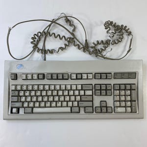 Puede incluir: Un teclado de ordenador IBM vintage con un cuerpo gris claro y teclas blancas. El teclado tiene una distribución QWERTY estándar con un teclado numérico y teclas de función. Un cable gris en espiral se extiende desde la parte superior del teclado.