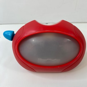 Pode incluir: Um visor View-Master vermelho com uma alavanca azul. O visor tem uma janela de visualização oval e as palavras "View-Master" estão impressas na frente. O dispositivo é feito de plástico e foi projetado para visualizar imagens 3D.