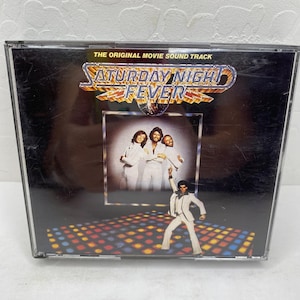 Puede incluir: Una caja de CD de la banda sonora de la película "Saturday Night Fever". La portada presenta el título en dorado y un diseño de bola de discoteca. La imagen incluye una foto de los Bee Gees y un hombre con traje blanco.