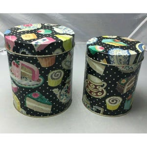 May include: Two round metal canisters with black and white polka dot backgrounds. The canisters are decorated with colourful images of cupcakes, cakes, teacups, and teapots.