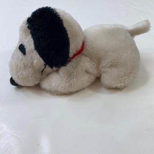 May include: A plush toy dog in a lying position. The dog has a white body, black ears, and a black nose. It wears a red collar. The toy is made of soft, fluffy material.