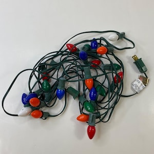 May include: A tangled string of Christmas lights with various colored bulbs, including red, blue, green, orange, and white. The lights are connected by a green wire and have a standard plug.