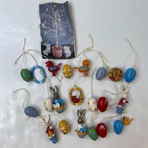 Pode incluir: Uma coleção de enfeites de Páscoa, incluindo ovos pintados, coelhos e pássaros. Os enfeites são em várias cores, como vermelho, azul, amarelo e verde, e são amarrados em barbante. Uma impressão vintage de uma árvore com ovos de Páscoa também está incluída.