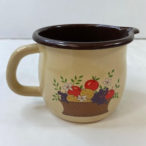 Op de afbeelding: Crèmekleurige emaille kan met een bruine rand en handvat. De kan heeft een fruitmandontwerp met rode appels, paarse druiven en witte bloemen. De kan heeft een tuit om gemakkelijk te schenken.