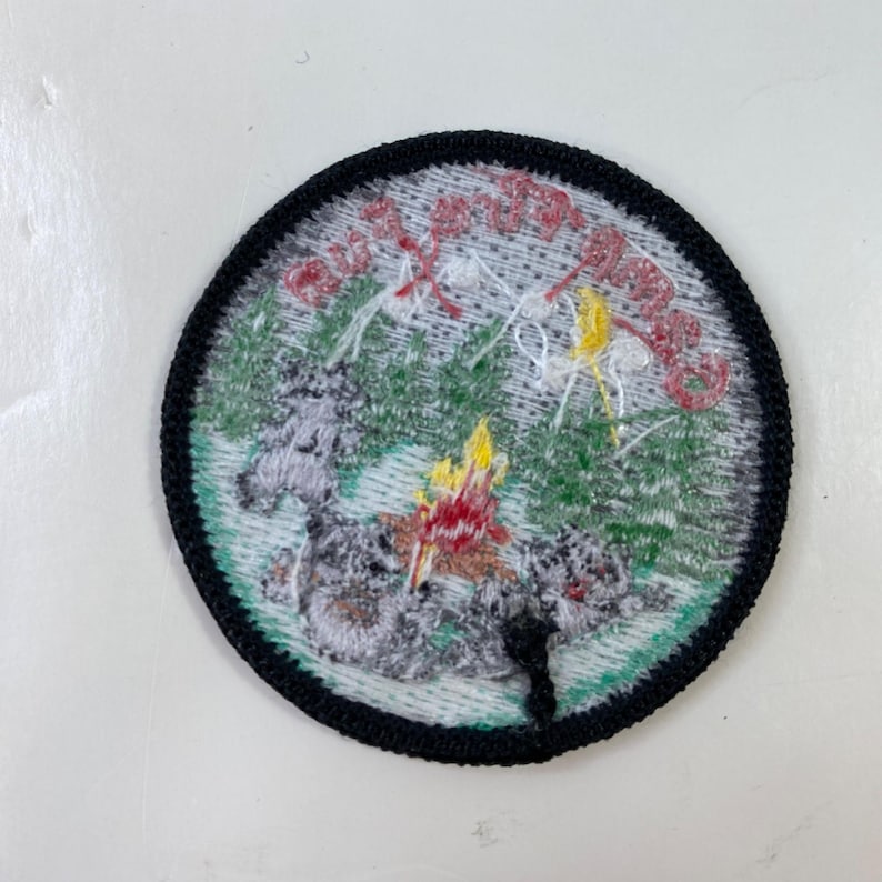 May include: Round embroidered patch with a black border. The patch features a campfire scene with trees, a mountain, and text in red. The patch is primarily green, gray, yellow, and red.