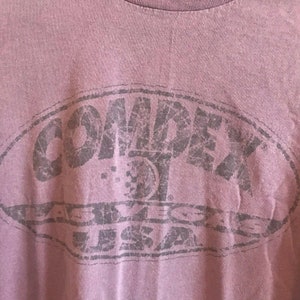 May include: A purple t-shirt with a distressed black graphic of the word "COMDEX" in a circle with the text "ABBEVILLE USA" underneath.