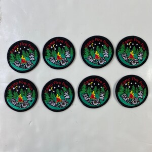 May include: Eight circular embroidered patches with a camping theme. Each patch features a campfire, raccoons, trees, stars, and the text "Camp Fire Fun" in a circular design. The patches have a black border.