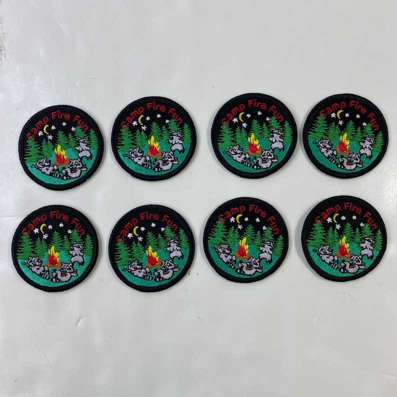 May include: Eight circular embroidered patches with a camping theme. Each patch features a campfire, raccoons, trees, stars, and the text "Camp Fire Fun" in a circular design. The patches have a black border.