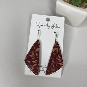 Puede incluir: Un par de pendientes de corcho burdeos y beige con un diseño de hojas. Los pendientes triangulares cuelgan de ganchos de metal oscuro. Se muestran en una tarjeta blanca con el texto "Spice by Jules".