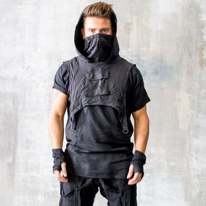 Op de afbeelding: Zwart ensemble bestaande uit een vest met capuchon, bijpassende shorts en een shirt met korte mouwen. Het vest heeft twee voorzakken en een gedrapeerd ontwerp. De outfit bevat vingerloze handschoenen en hoge sneakers met bandjes. Het model staat voor een grijze muur.