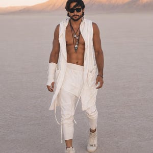 Könnte beinhalten: Eine Person trägt eine weiße ärmellose Kapuzenweste, eine passende Hose und High-Top-Sneakers. Das Outfit umfasst mehrere Halsketten und eine Sonnenbrille. Der Hintergrund zeigt eine Wüstenlandschaft mit Bergen unter einem warm getönten Himmel.