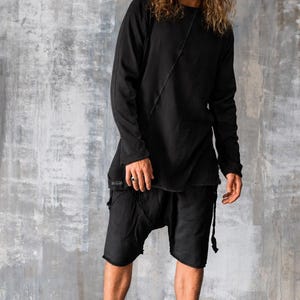 May include: Black long-sleeve top and shorts outfit. The top has a diagonal seam detail and a slightly asymmetrical hem. The shorts have a drop-crotch design and frayed edges. The outfit is paired with black high-top sneakers.