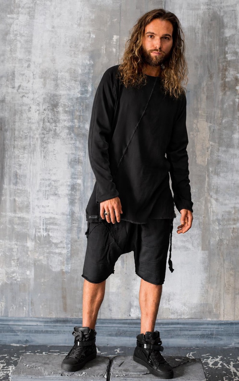 May include: Black long-sleeve top and shorts outfit. The top has a diagonal seam detail and a slightly asymmetrical hem. The shorts have a drop-crotch design and frayed edges. The outfit is paired with black high-top sneakers.