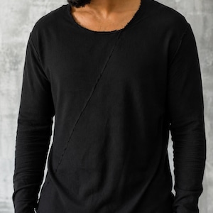 May include: Black long-sleeve shirt with a raw-edge neckline and a diagonal seam across the front. The shirt has long sleeves and a relaxed fit. The fabric appears to be a soft, lightweight material. The shirt is a solid black color.