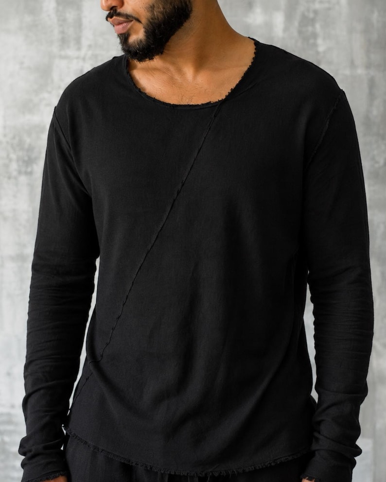 May include: Black long-sleeve shirt with a raw-edge neckline and a diagonal seam across the front. The shirt has long sleeves and a relaxed fit. The fabric appears to be a soft, lightweight material. The shirt is a solid black color.