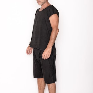 May include: A black linen outfit consisting of a short-sleeved top and matching shorts. The outfit has a relaxed fit and a textured appearance. The model is standing against a white background, showcasing the casual style of the clothing.