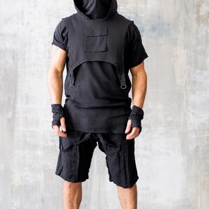 May include: Black hooded vest with a pocket, worn over a black t-shirt. The outfit includes black shorts, fingerless gloves, and high-top sneakers. The clothing has a rugged, urban aesthetic.