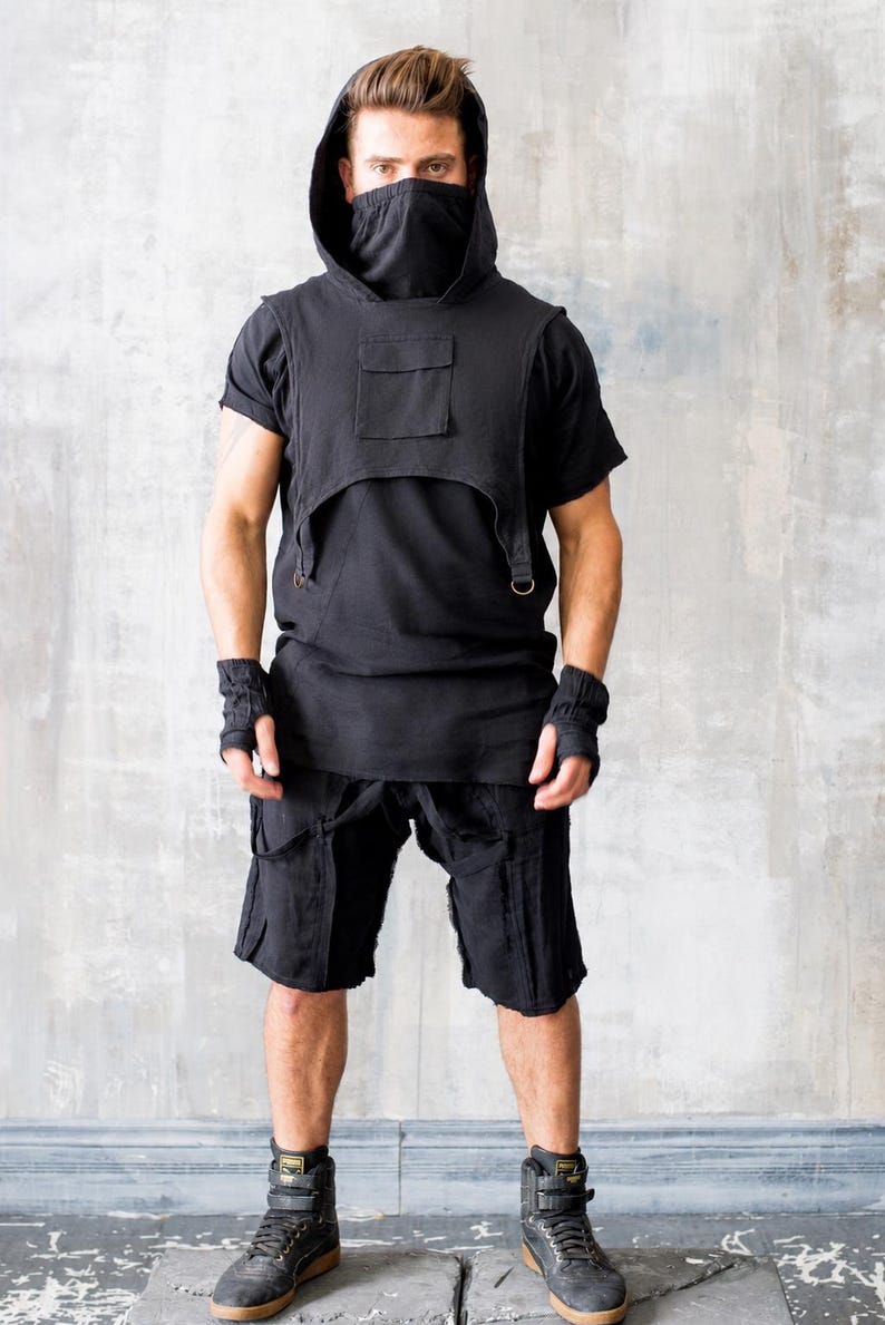 May include: Black hooded vest with a pocket, worn over a black t-shirt. The outfit includes black shorts, fingerless gloves, and high-top sneakers. The clothing has a rugged, urban aesthetic.