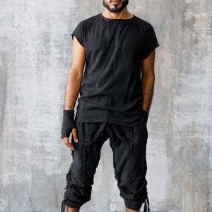 May include: Black outfit consisting of a short-sleeved top and cropped pants. The pants have multiple pockets and straps. The outfit is paired with black high-top sneakers. The person is standing in front of a gray wall.