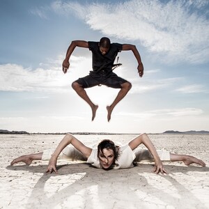 May include: Two people performing dance moves outdoors. One person is in mid-air, wearing a black outfit. The other person is on the ground, in a split, wearing a white outfit. The background is a dry, cracked landscape under a blue sky.