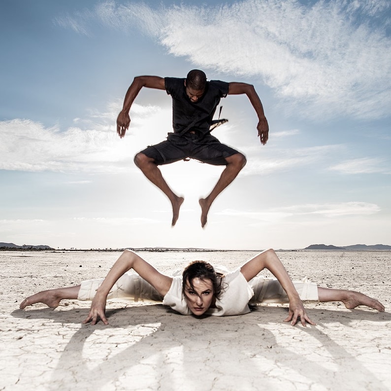 May include: Two people performing dance moves outdoors. One person is in mid-air, wearing a black outfit. The other person is on the ground, in a split, wearing a white outfit. The background is a dry, cracked landscape under a blue sky.