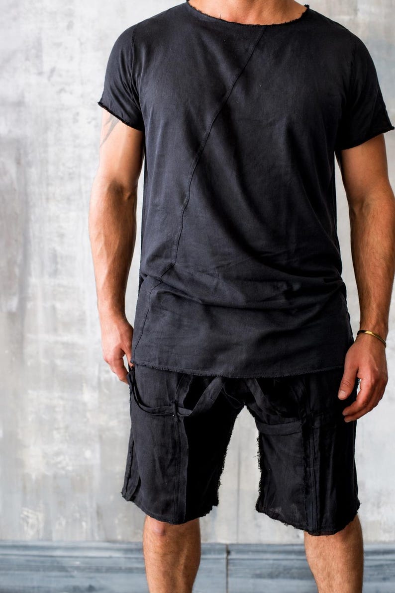 May include: Black t-shirt and shorts set. The short-sleeved shirt has a raw edge neckline and a diagonal seam. The shorts have straps and pockets. The outfit is made of a soft, dark fabric.