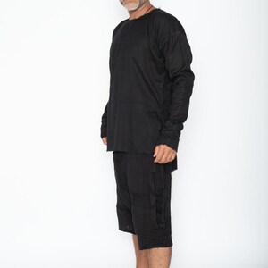 May include: Black long-sleeve shirt and matching shorts. The shirt has a crew neck and long sleeves. The shorts are knee-length with pockets. The outfit is made of a textured fabric. The person is standing barefoot against a white background.