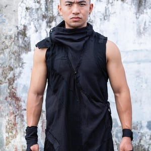 May include: A person wearing a black sleeveless shirt with a high collar and a zipper. The shirt has a distressed look with frayed edges. The person is standing in front of a concrete wall.