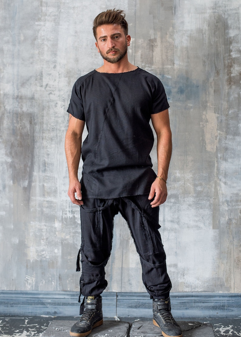 May include: A person wearing a black short-sleeved shirt and black pants. The shirt has a unique neckline and a diagonal seam. The pants feature multiple pockets and straps. The person is wearing black high-top sneakers.