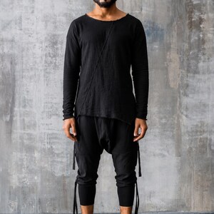 May include: A person wearing a black long-sleeved top with a diagonal seam and matching black pants. The pants have attached straps and are paired with white high-top sneakers.