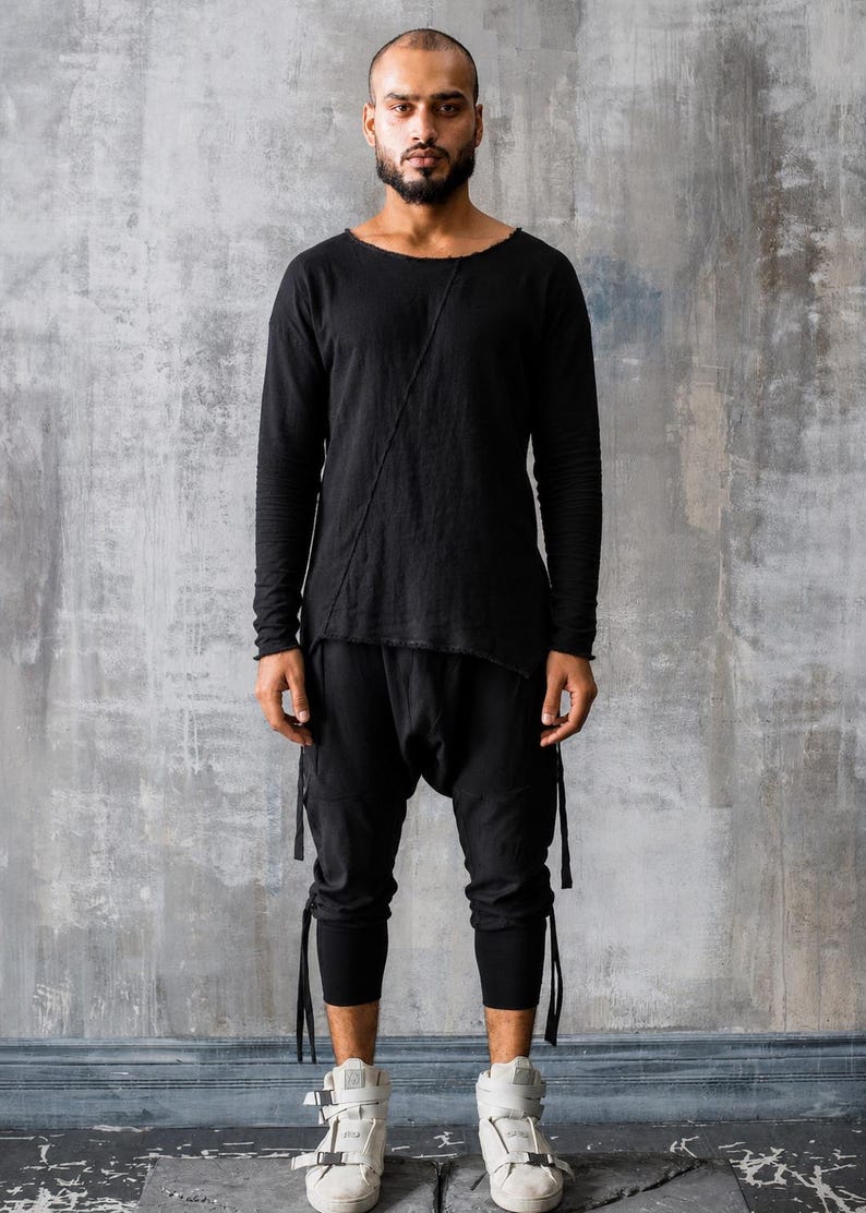 May include: A person wearing a black long-sleeved top with a diagonal seam and matching black pants. The pants have attached straps and are paired with white high-top sneakers.