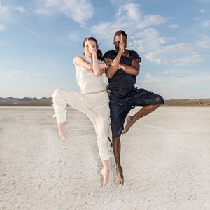 May include: Two people in athletic attire are posed in a balanced stance against a backdrop of a dry, cracked landscape under a blue sky. One person wears white, the other black. Both have their hands in a prayer position.