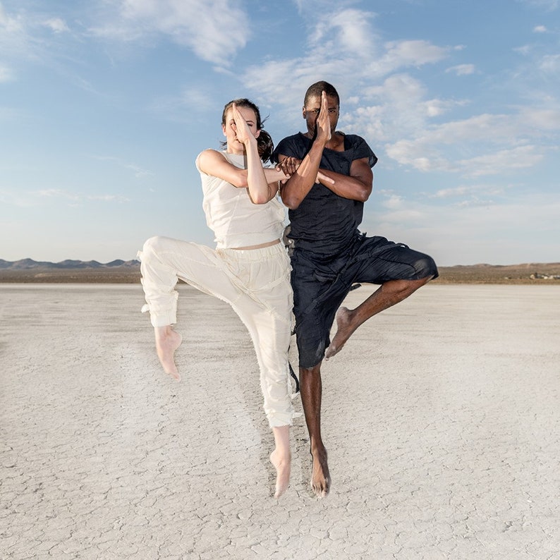 May include: Two people in athletic attire are posed in a balanced stance against a backdrop of a dry, cracked landscape under a blue sky. One person wears white, the other black. Both have their hands in a prayer position.