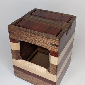 May include: A wooden box with a square shape and a rectangular opening in the front. The box is made of different types of wood, including walnut, maple, and cherry. The wood is arranged in a striped pattern.