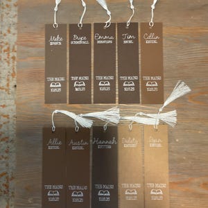 Può includere: Una collezione di segnalibri personalizzati con nappe bianche. Ogni segnalibro presenta un nome, il testo "THE MAINS" e la data "10.18.25" o "10.15.25". I segnalibri sono in varie tonalità di marrone e beige.