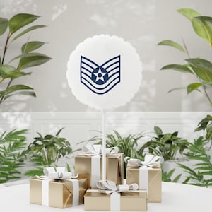 May include: A white, round balloon with a navy blue military insignia, hovering above several gold gift boxes tied with white ribbons. The gifts are arranged on a white surface, with green plants in the background.