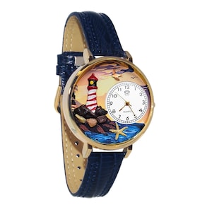 Könnte beinhalten: Goldfarbene Armbanduhr mit marineblauem Lederarmband. Das Zifferblatt zeigt eine Miniatur-Leuchtturmszene mit einem rot-weiß gestreiften Leuchtturm, Felsen und einem Seestern. Die Uhr hat ein weißes Zifferblatt mit schwarzen Ziffern und Zeigern.