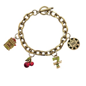 May include: Gold-toned charm bracelet featuring casino-themed charms. Charms include a slot machine, cherries, a lucky number seven, and a casino chip with "$100 Casino" text. The bracelet has a toggle clasp.