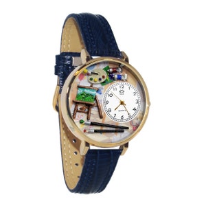May include: Gold-tone quartz watch with a blue leather band. The watch face features a miniature artist's easel, paint tubes, palette, and brushes. The watch has a white face with black numerals and hands.