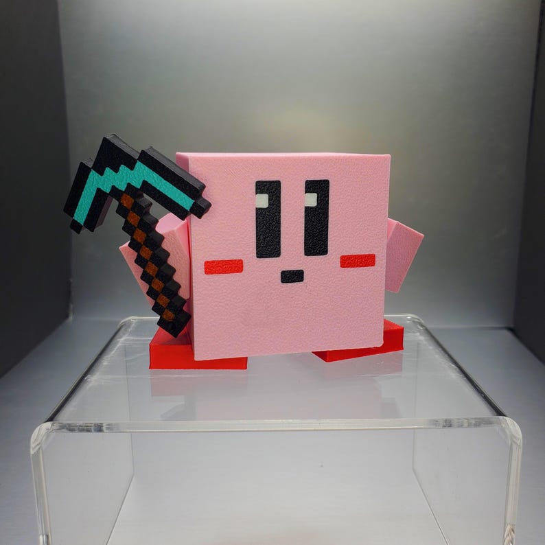 May include: A pink, pixelated cube-shaped character with black eyes and a small mouth, holding a pixelated pickaxe. The character has small red feet and cheeks. The item is a 3D-printed figurine.