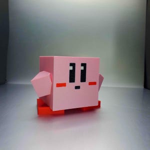 May include: A pink, cube-shaped figure with black eyes and mouth, and red accents. The figure has small, pink, rectangular arms and is standing on a red base. The design is reminiscent of a popular video game character.